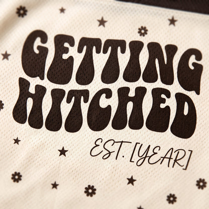 Close-up of a cream-colored mesh fabric with dark brown retro-style text reading 'GETTING HITCHED' and smaller script 'EST. [YEAR]', decorated with small star and flower motifs.