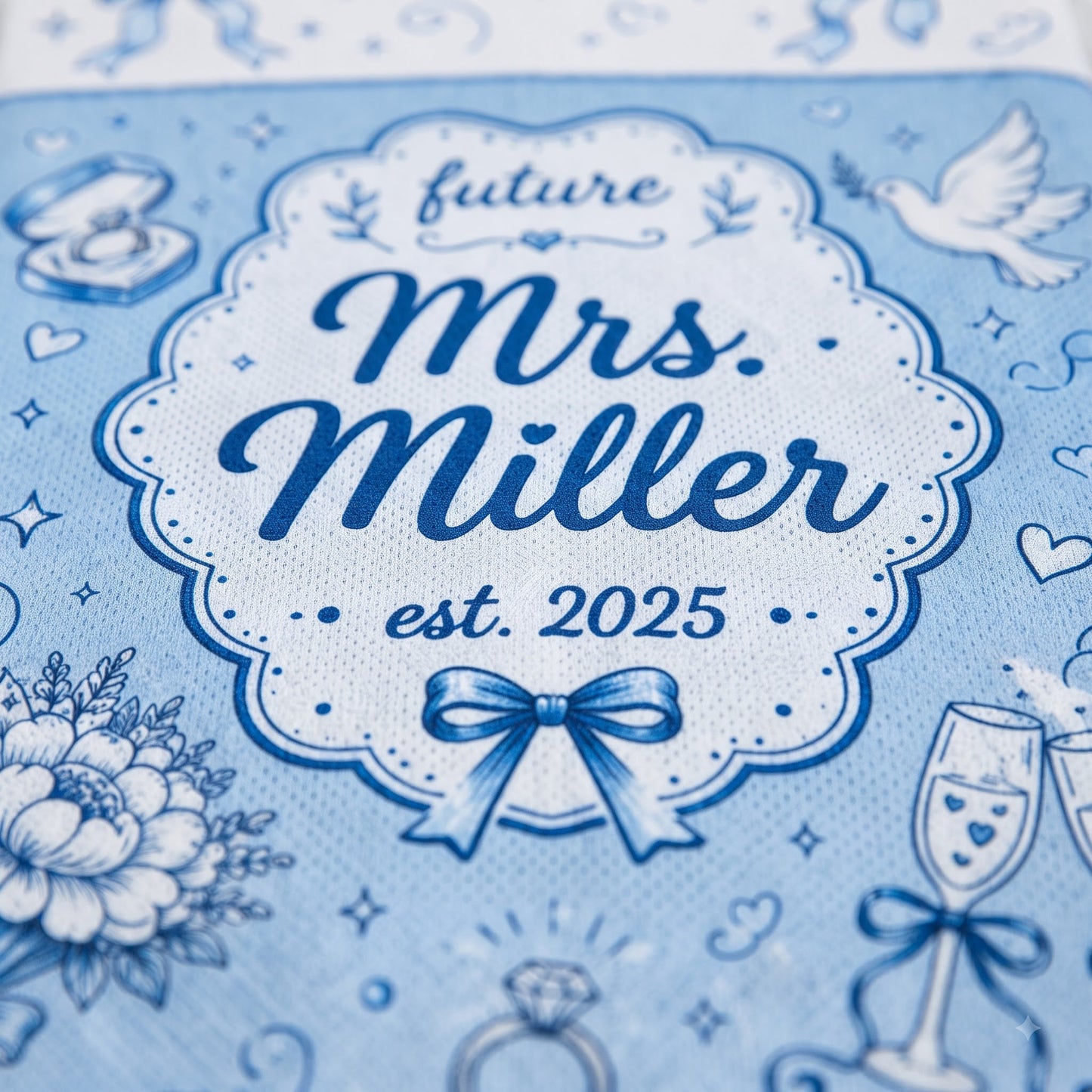 A close-up of a light blue fabric item printed with a decorative white and navy label reading 'future Mrs. Miller est.