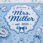 A close-up of a light blue fabric item printed with a decorative white and navy label reading 'future Mrs. Miller est.