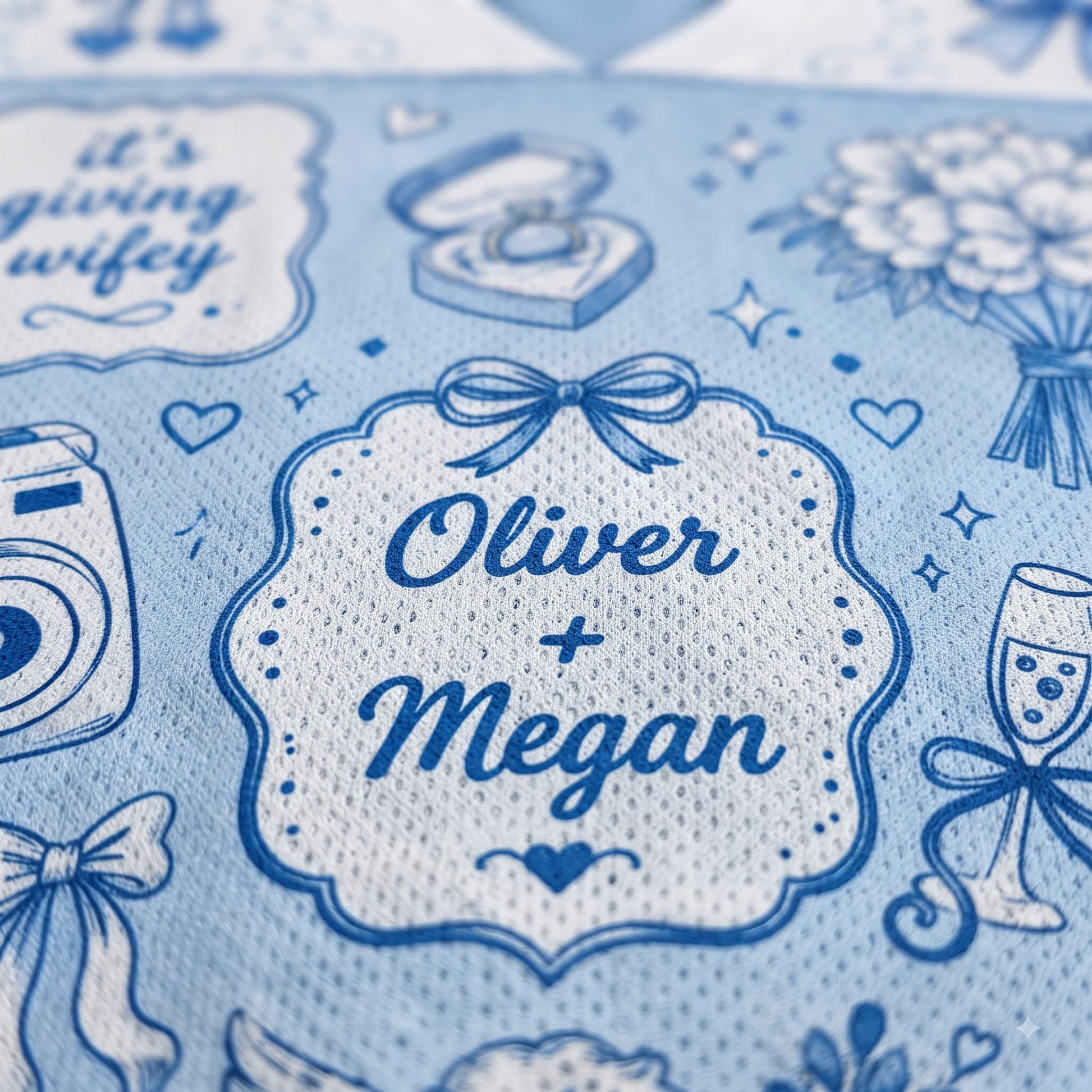 Close-up of a personalized light blue fabric item, likely a gift wrap or banner, featuring a central white scalloped label with the names 'Oliver + Megan' in blue script. The fabric is decorated with blue wedding-themed illustrations including a ring box, bouquet, champagne glass, and camera, alongside the phrase 'it's giving wifey'.