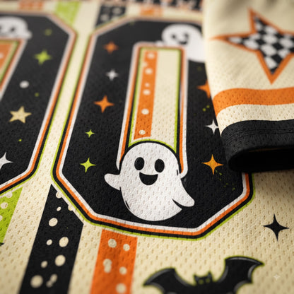 Close-up of a Halloween-themed sports jersey fabric featuring a black number '0' with a white cartoon ghost, orange stars, green accents, and a cream mesh background.