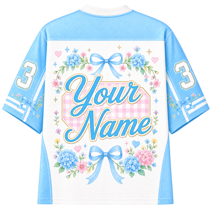 A customizable short-sleeve athletic jersey shown from the back, featuring a white center panel with a pink gingham background, large script text reading 'Your Name,' blue floral arrangements, bows, stars, and the number 3 on both sleeves.