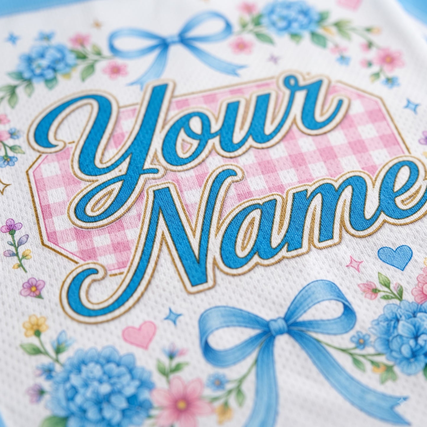 Close-up of a personalized white fabric item featuring blue script text reading 'Your Name' over a pink gingham shape, decorated with blue bows, blue flowers, and small pink and blue floral accents.