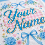 Close-up of a personalized white fabric item featuring blue script text reading 'Your Name' over a pink gingham shape, decorated with blue bows, blue flowers, and small pink and blue floral accents.