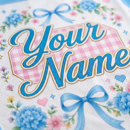 Close-up of a personalized white fabric item featuring blue script text reading 'Your Name' over a pink gingham shape, decorated with blue bows, blue flowers, and small pink and blue floral accents.