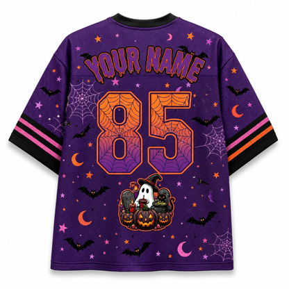 A purple short-sleeve t-shirt with a Halloween theme, featuring customizable text 'YOUR NAME' and the number '85' on the back. The design includes orange spiderwebs, black bats, pink and orange crescent moons, purple stars, spiderwebs, and a cartoon ghost and pumpkin decoration near the hem.