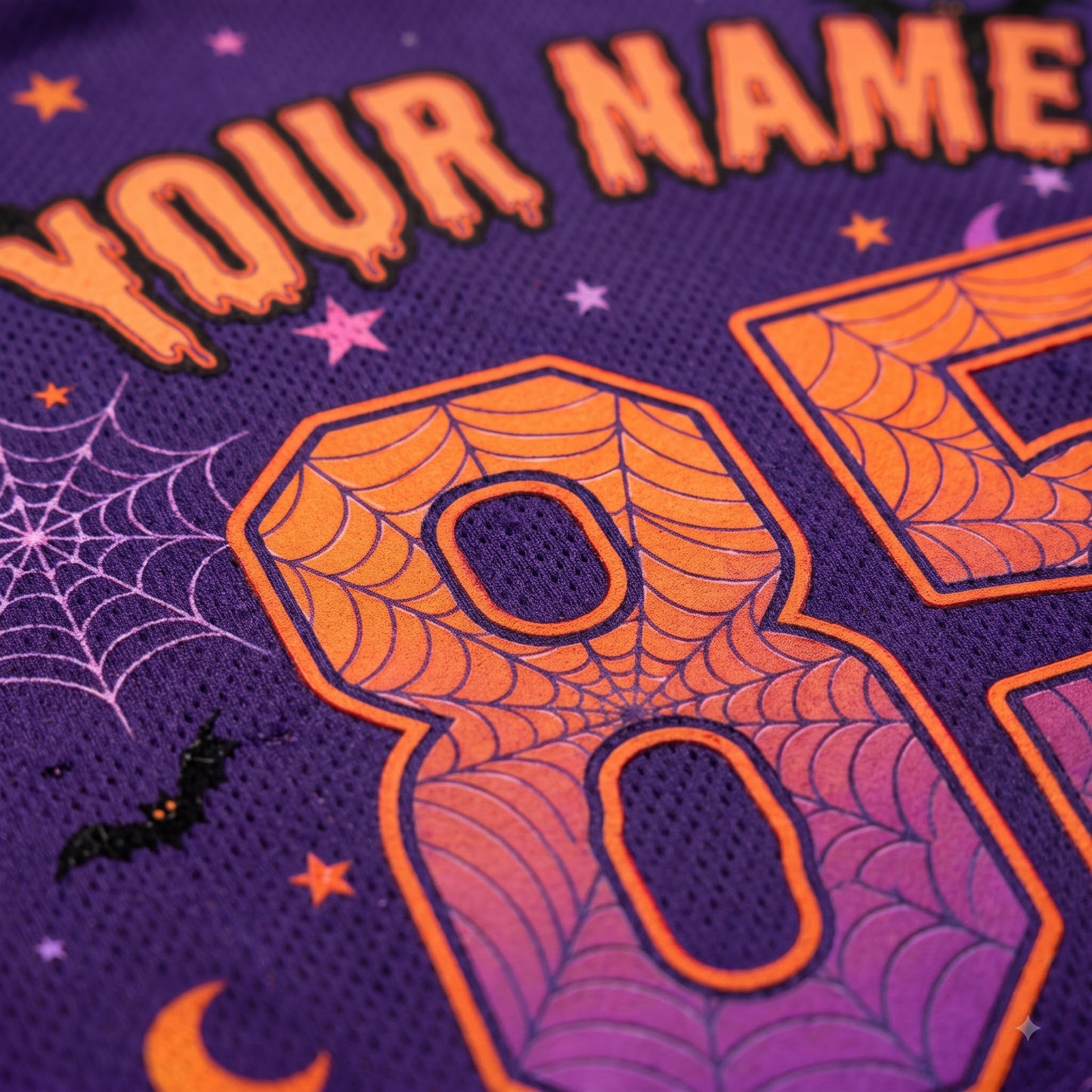 Close-up of a purple mesh fabric with orange and pink Halloween-themed embroidery, including a large spiderweb-style number 8, partial 'YOUR NAME' text, stars, web motifs, and a small bat.