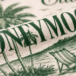 Close-up of a rolled fabric or napkin with a tropical beach print. The design features dark green text reading 'HONEYMOON' alongside illustrations of palm trees, a beach chair, and ocean scenery on a warm cream background.