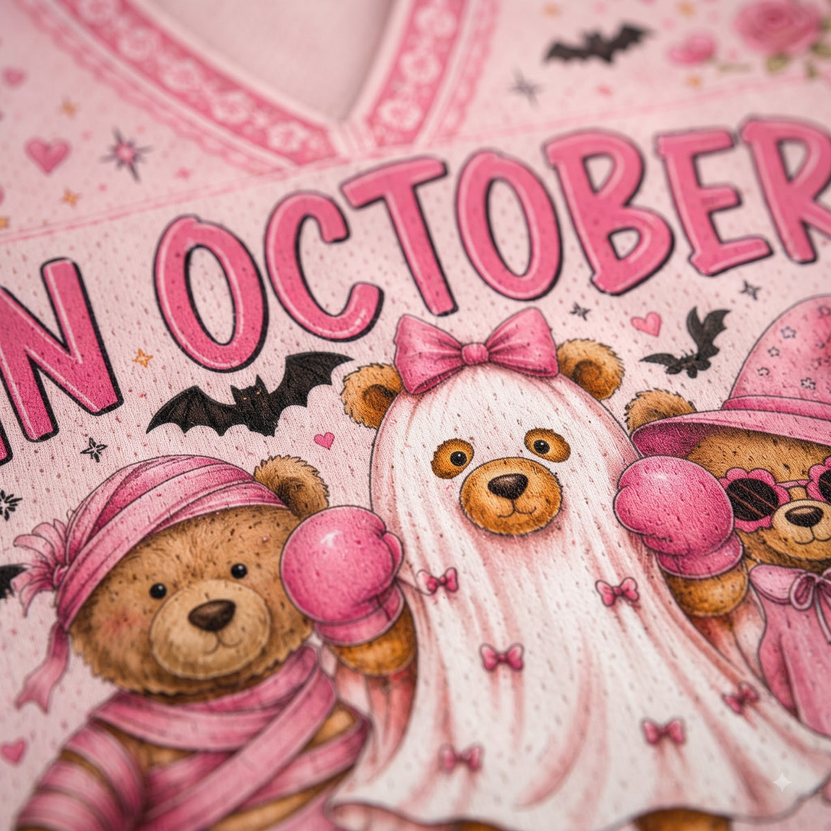 Close-up of a pink Halloween-themed garment featuring illustrated teddy bears in costumes with the text 'IN OCTOBER' and small bat and heart motifs.
