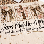 Close-up of a cream-colored garment featuring a graphic print with a wooden cross draped in pink fabric, leopard-print bows, roses, and the text 'Jesus Made Her A Mom and she wouldn't have it any other way'.