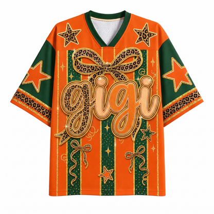 An orange and green graphic t-shirt with a leopard print bow, stars, and the word 'gigi' in large gold script on the front, displayed on a clean white background.