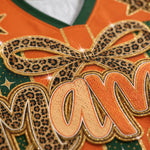 Close-up of an embroidered sports jersey detail featuring leopard-print applique, orange mesh lettering outlined in glittery gold, and green trim.