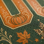 Close-up of a dark green textured fabric featuring a large orange letter U with a pumpkin motif, decorated with gold-toned scrollwork, an orange maple leaf, and small stars.