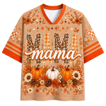 A short-sleeve V-neck graphic shirt in a warm beige tone with orange trim, featuring the word 'MAMA' in large patterned letters and an autumn floral and pumpkin print. The sleeves have orange stripes and patterned accents.