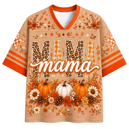 A short-sleeve V-neck graphic shirt in a warm beige tone with orange trim, featuring the word 'MAMA' in large patterned letters and an autumn floral and pumpkin print. The sleeves have orange stripes and patterned accents.
