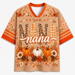 A short-sleeve V-neck t-shirt with a beige mesh-like background, featuring a festive autumn theme. The design includes the word 'NANA' in large leopard-print and striped letters, a white script 'nana' across the center, orange pumpkins, white and orange flowers, pinecones, and orange leaves.