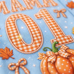 A close-up of a light blue fabric surface with large orange-and-white gingham numbers 0 and 1, autumn leaves, bows, and a pumpkin decoration.