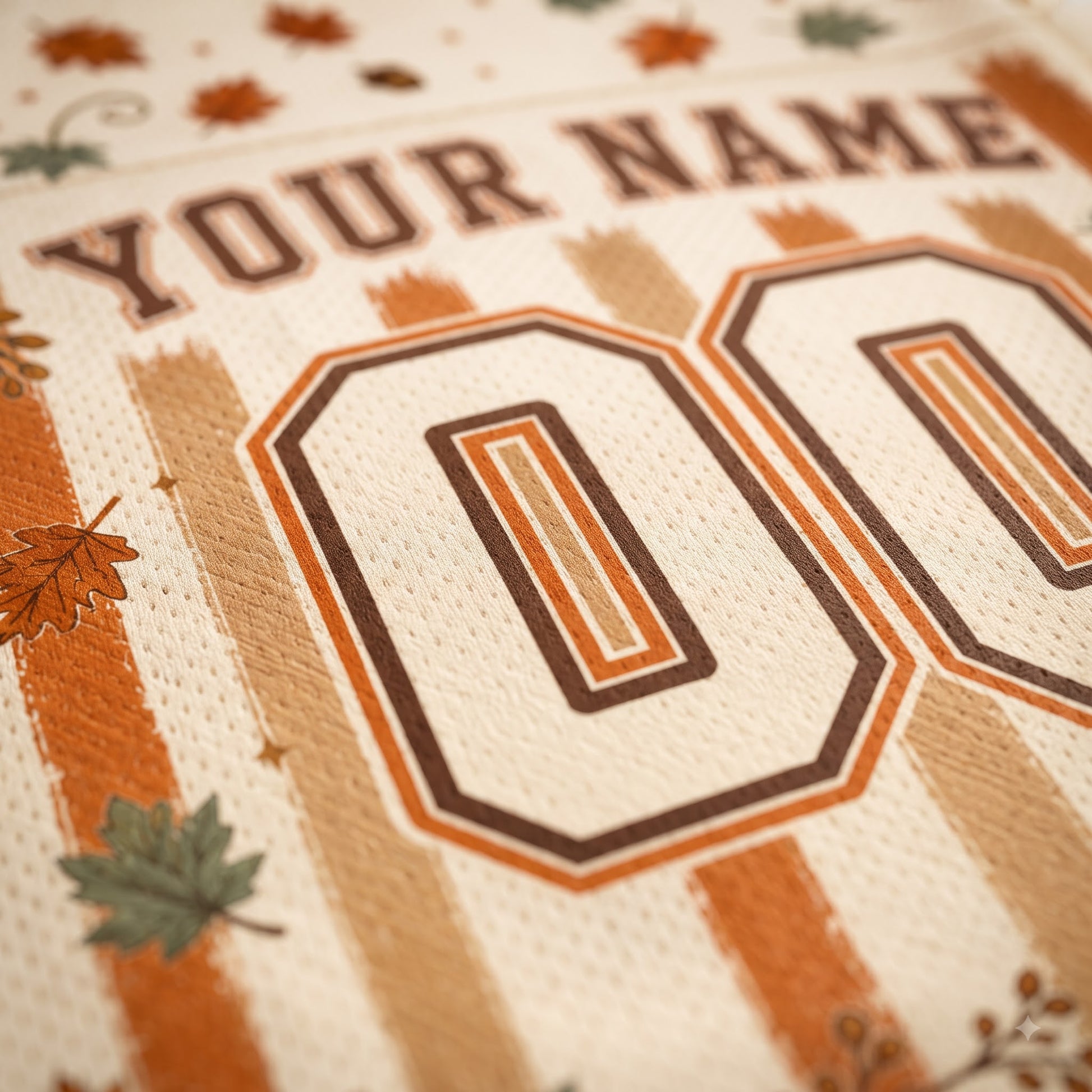 Close-up of a personalized sports jersey fabric with the text 'YOUR NAME' and large '00' numbers in brown and orange, decorated with autumn leaves and stripes on a cream background.