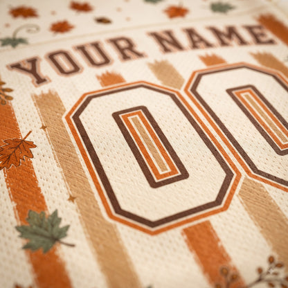 Close-up of a personalized sports jersey fabric with the text 'YOUR NAME' and large '00' numbers in brown and orange, decorated with autumn leaves and stripes on a cream background.