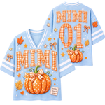 A light blue short-sleeve V-neck jersey-style top shown from both front and back, decorated with orange gingham 'MIMI' lettering, large orange pumpkin graphics with bows, white stars, fall leaves, and white sleeve stripes. The back also shows 'MIMI 01' in orange gingham.