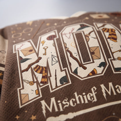 Close-up of a brown knit garment featuring large decorative letters filled with whimsical illustrations and the text 'Mischief Ma.