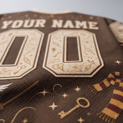 Close-up of a dark brown personalized sports jersey with cream-colored custom number 00 and 'YOUR NAME' text, decorated with Harry Potter themed illustrations including keys, wand parts, stars, and a striped scarlet scarf.
