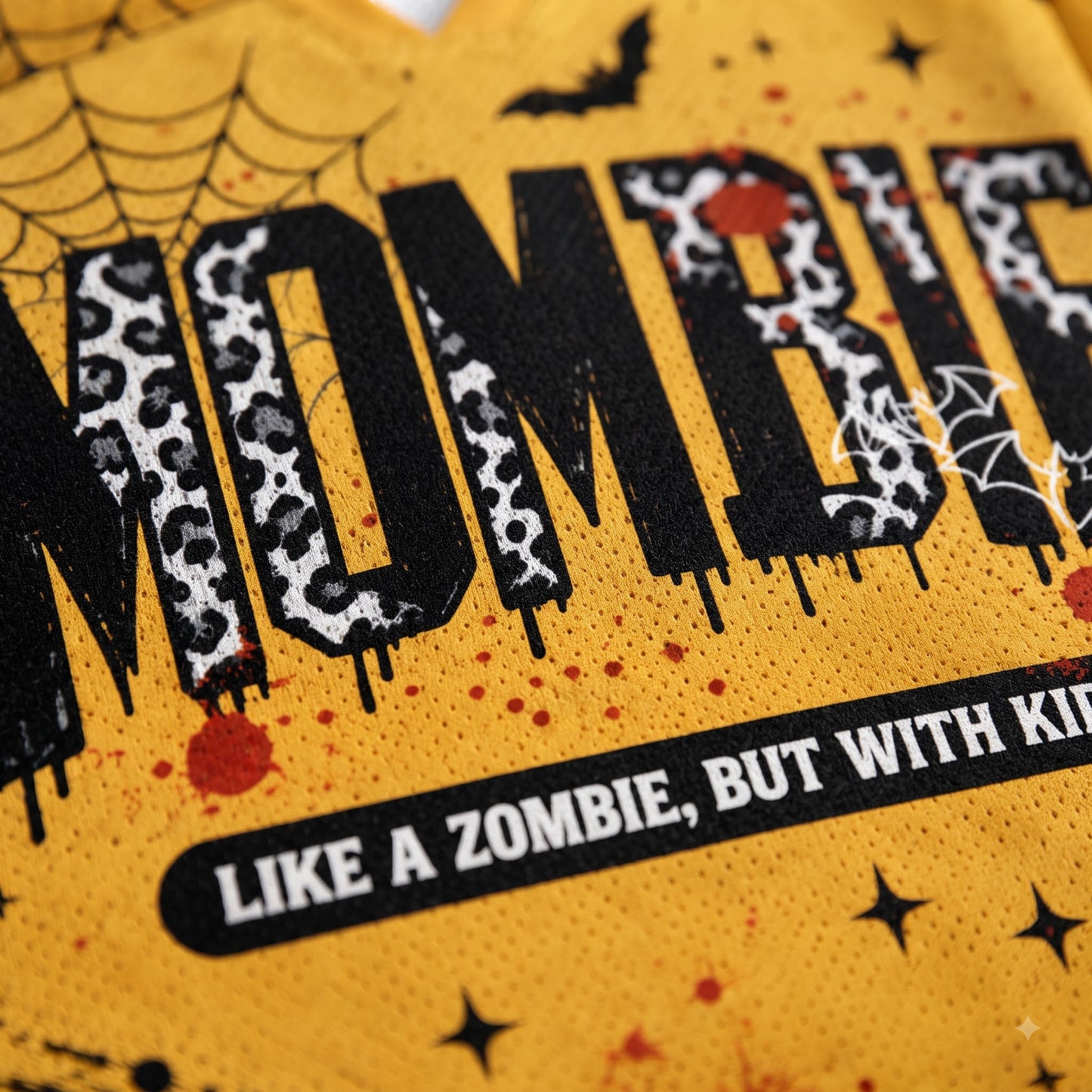 Close-up of a yellow graphic print featuring the word 'MOMMY' in large black letters with a black-and-white leopard print pattern, surrounded by Halloween-themed graphics including spiderwebs, stars, bats, and red splatter dots. Text below reads 'LIKE A ZOMBIE, BUT WITH KIDS'.