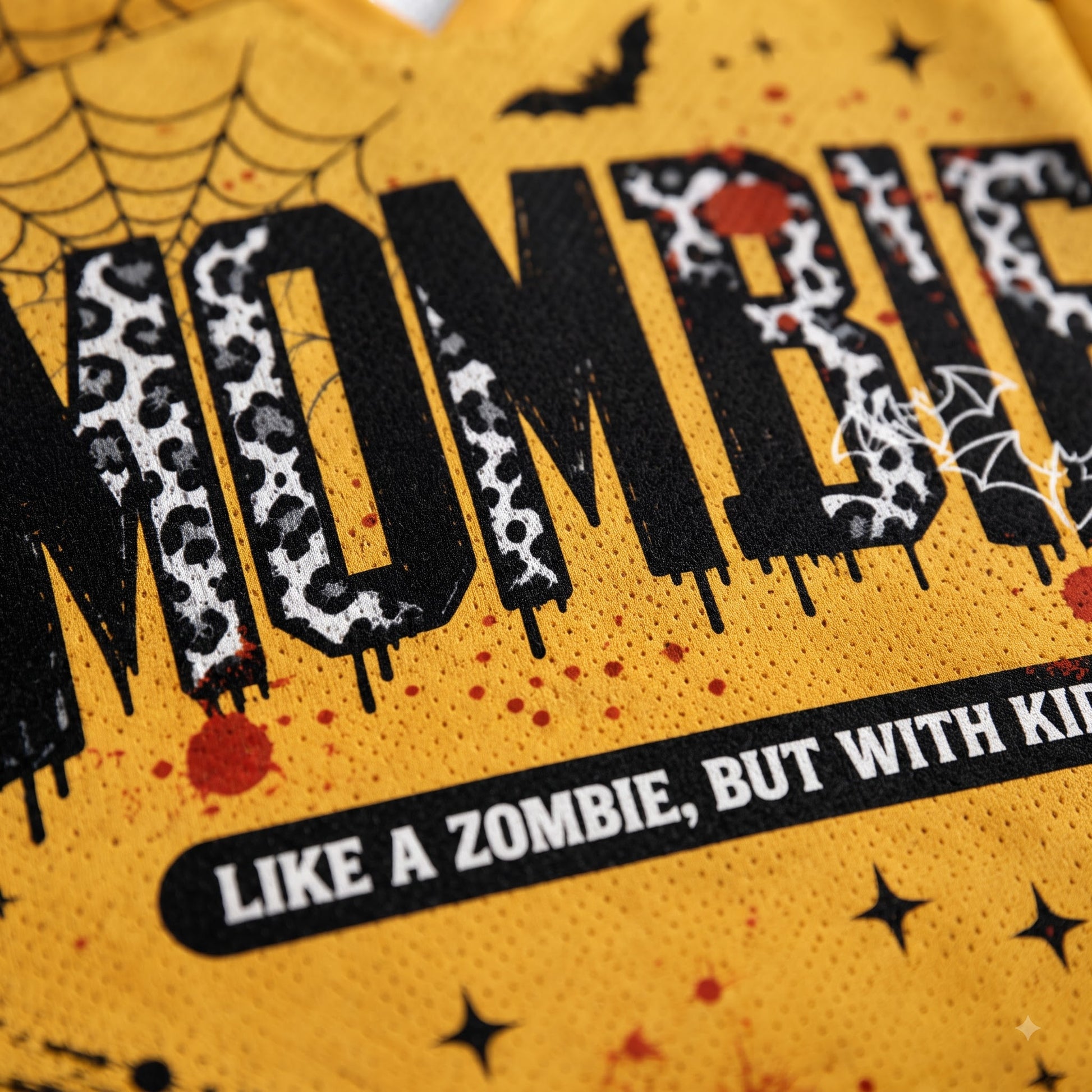 Close-up of a yellow graphic print featuring the word 'MOMMY' in large black letters with a black-and-white leopard print pattern, surrounded by Halloween-themed graphics including spiderwebs, stars, bats, and red splatter dots. Text below reads 'LIKE A ZOMBIE, BUT WITH KIDS'.