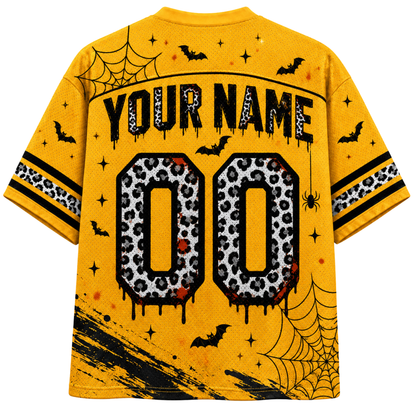 A yellow short-sleeve t-shirt shown from the back, featuring a Halloween-themed graphic design with 'YOUR NAME' and '00' in a black and white leopard print, decorated with black bats, spiderwebs, stars, and black paint splatters.