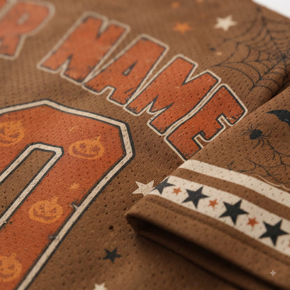 Close-up of a brown mesh sports jersey with Halloween-themed graphics, including orange outlined lettering, pumpkin motifs, spiderweb print, and black stars on a folded cuff.