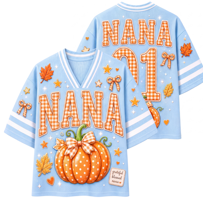 A light blue V-neck short-sleeve jersey-style shirt shown from both front and back, decorated with autumn graphics including a large orange pumpkin with a gingham bow, fall leaves, stars, and the word “NANA” in orange gingham lettering. The back also shows “NANA 91” in large numbers.