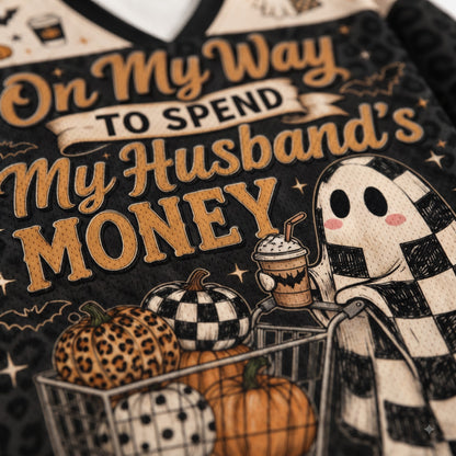 Close-up of a Halloween-themed graphic print on a garment, featuring the phrase 'On My Way TO SPEND My Husband's MONEY' in tan lettering over a black background, with pumpkins, a ghost in a checkered blanket, and a shopping cart.
