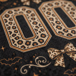 Close-up of a black patterned fabric featuring large decorative '0' numerals in a leopard print, a matching bow motif, and small gold-toned decorative accents.