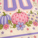 Close-up of a printed fabric or paper napkin featuring three decorative pumpkins in purple, pink, and white with green leaves, small pink and purple flowers, and a large lavender number 00 at the top.