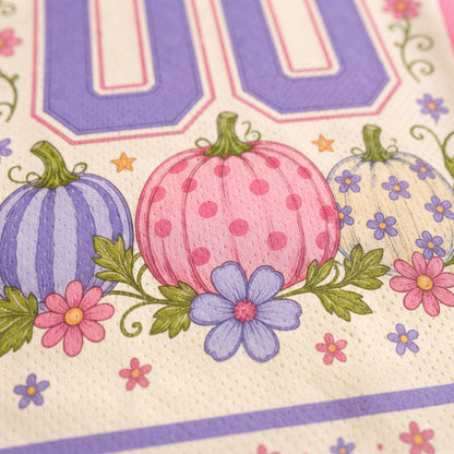 Close-up of a printed fabric or paper napkin featuring three decorative pumpkins in purple, pink, and white with green leaves, small pink and purple flowers, and a large lavender number 00 at the top.