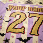 Close-up of a personalized sports jersey fabric with a purple and cream checkered pattern, featuring a large gold mesh number '27' with red and black outlines, and 'your name' printed above. The design includes black bats, stars, and pink accents.