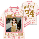 A personalized sports jersey shown from both front and back. The light pink jersey features pink and white striped trim, gold glitter-style text reading 'LOOK WHO'S GETTING MARRIED' and 'SOPHIA', a large gold question mark with '34', pink bows, hearts, flowers, champagne glasses, and a custom photo frame on the front.