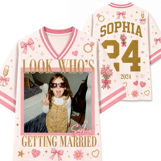 A personalized sports jersey shown from both front and back. The light pink jersey features pink and white striped trim, gold glitter-style text reading 'LOOK WHO'S GETTING MARRIED' and 'SOPHIA', a large gold question mark with '34', pink bows, hearts, flowers, champagne glasses, and a custom photo frame on the front.