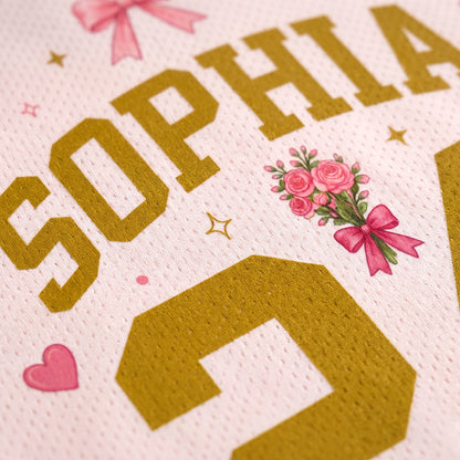 Close-up of a personalized sports jersey fabric featuring the name 'SOPHIA' in gold block letters, pink roses with a bow, and gold stars on a textured white background.
