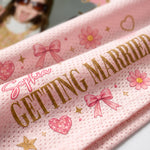 A close-up of a pink textured fabric item printed with the words 'GETTING MARRIED' in gold, along with pink bows, flowers, hearts, and the name 'Sophia' in pink script.