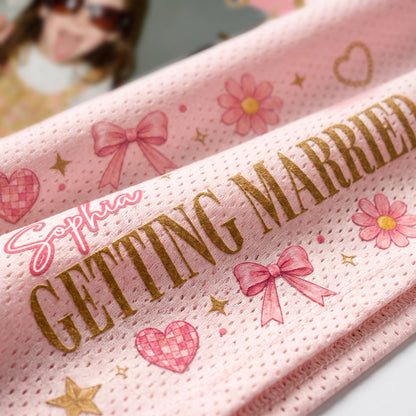 A close-up of a pink textured fabric item printed with the words 'GETTING MARRIED' in gold, along with pink bows, flowers, hearts, and the name 'Sophia' in pink script.