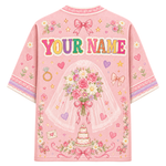 A pink short-sleeve t-shirt with a customizable graphic design on the back, featuring the text 'YOUR NAME' in colorful letters, a bride holding a rose bouquet under a veil, a wedding cake, and various floral and heart motifs.