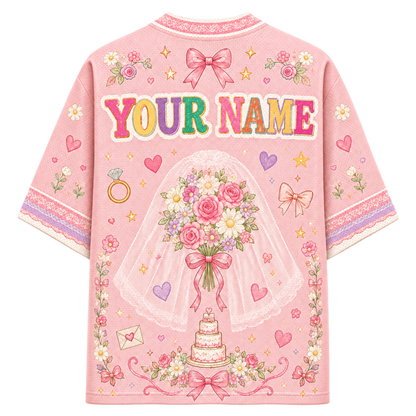 A pink short-sleeve t-shirt with a customizable graphic design on the back, featuring the text 'YOUR NAME' in colorful letters, a bride holding a rose bouquet under a veil, a wedding cake, and various floral and heart motifs.