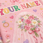 A close-up of a pink personalized wedding napkin or paper plate featuring a bride bouquet illustration, a ring, stars, hearts, and the text 'YOUR NAME' in colorful letters.