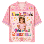 A pink short-sleeve V-neck t-shirt featuring a colorful graphic print of a young girl in a floral crown with the text 'Look Who's Getting Married' in multicolored letters, surrounded by hearts, stars, and floral motifs.