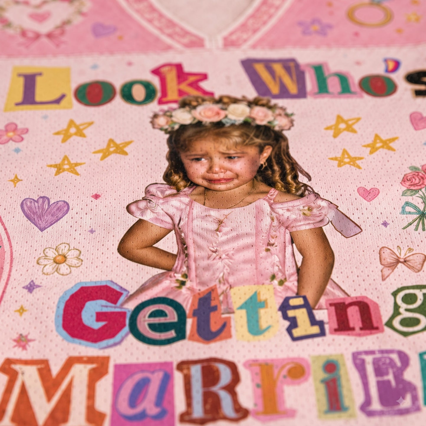 A close-up of a pink novelty blanket or fabric featuring a printed photo of a young girl in a pink dress and flower crown, surrounded by colorful text reading 'Look Who's Getting Married' and decorative stars, hearts, and flowers.