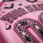 Close-up of a pink athletic jersey with black and pink rhinestone lettering, bat motifs, and a large black-and-pink number detail on the front.