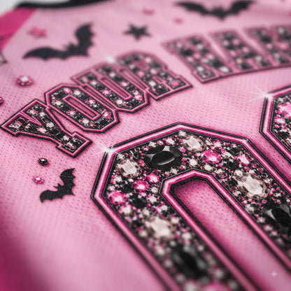 Close-up of a pink athletic jersey with black and pink rhinestone lettering, bat motifs, and a large black-and-pink number detail on the front.