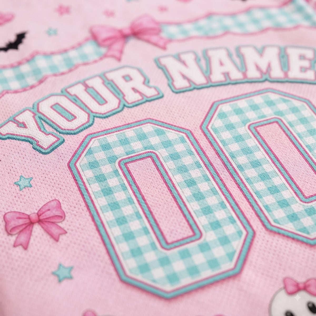 Close-up of a pink fabric item with embroidered text reading 'YOUR NAME' and large gingham-style numbers '00' in light blue and white, decorated with pink bows and small blue stars.