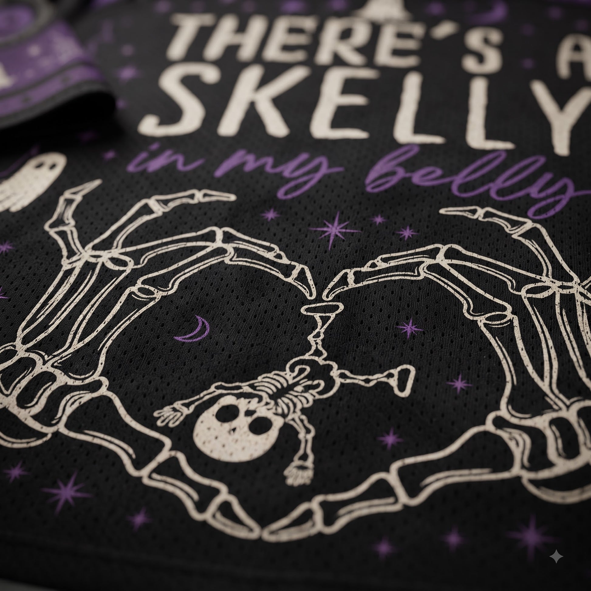 Close-up of a black mesh garment featuring a white skeleton graphic forming a heart shape around a small skull, with purple stars and the text 'THERE'S A SKELLY in my belly' in white and purple lettering.